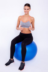 Staying fit. Young beautiful woman in sportswear with perfect bodie thumbs and looking at camera with smile while sitting on fitness balls over white isolated background.