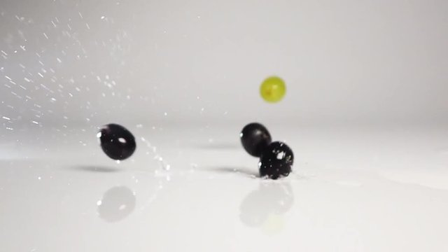 Fresh tasty green and black grapes berries dropping down and jump on water surface on wet white floor with stunning splashing and spraying. Shot with high speed camera in slow motion mode. White