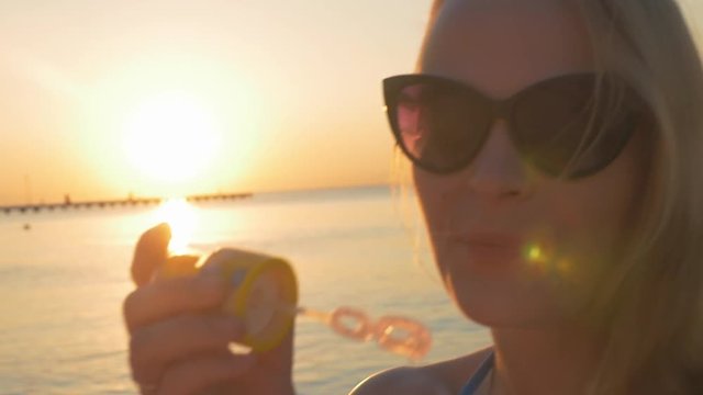 Slow motion shot of young blonde woman in sunglasses blowing soap bubbles at sunset.