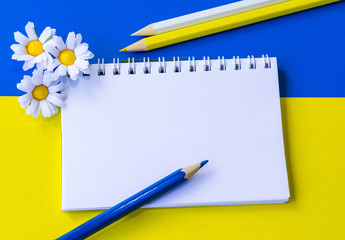 Blank notepad decorated by artificial camomiles and colorful pencils on the yellow and blue...