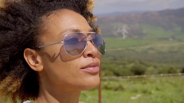 Thoughtful young woman wearing trendy sunglasses looking out over a rural landscape with a pensive expression  close up profile