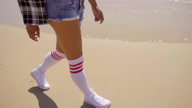 Close up of legs of young black woman as she walks a sandy beach along. She escapes the water waves and have lot of fun. The camera pans up from her feets to upper body 