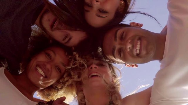 Low angle view of five good looking friends in a huddle as they smile with the sun beating down behind them