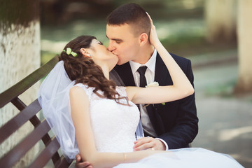 Attractive brunette bride in elegant dress and handsome groom in blue suit at wedding day lovely hugging on bench outdoors. Newlywed woman and man embracing in green park. Loving wedding couple