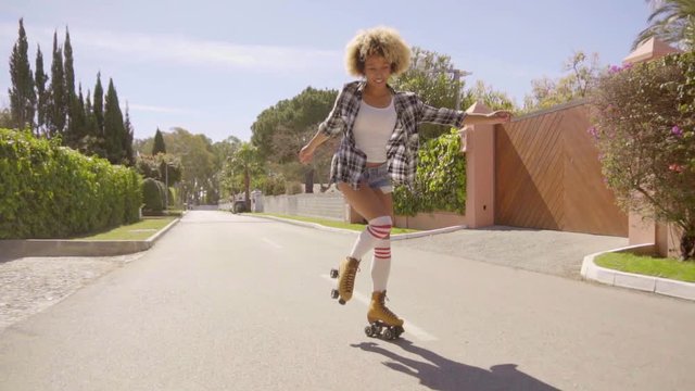 Slow motion view of young afro-american woman on vintage roller skates rides through the upmarket village streets and showing her driving skills.