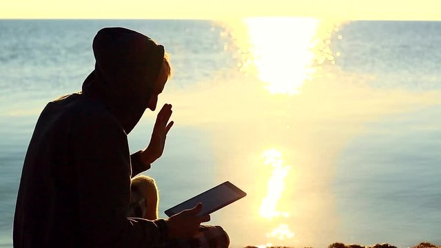 A guy with a tablet sitting on the gravel beach in front of the sea 