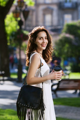 Portrait of young beautiful happy smiling lady walking on the street, holding paper cup of coffee. Model wearing stylish clothes & accessories. Girl looking at camera. City lifestyle. Sunny day. 