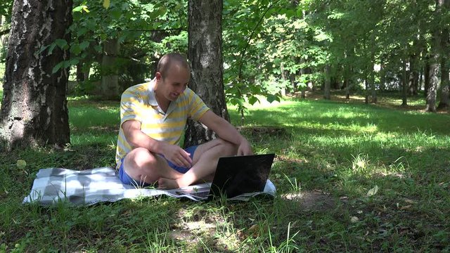 Happy man in front of computer screen. Smiling guy with laptop chatting with girlfriend online in park. Static shot. 4K
