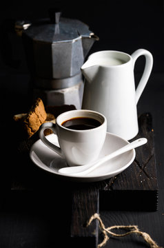 Cup of hot espresso, creamer with milk, cantucci and moka coffee pot on a rustic wooden board