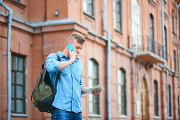 Fototapeta premium Student in a denim shirt, talking on a cell phone and holds your tablet in hand on the background of the building of brick
