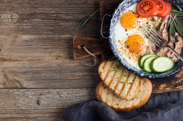 Breakfast set. Pan of fried eggs with bacon, fresh tomato, cucumber, sage and bread on rustic serving board over wooden background