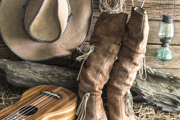 Closeup american west rodeo brown felt cowboy hat and traditional leather boots with ukulele in vintage ranch barn background, Still life style