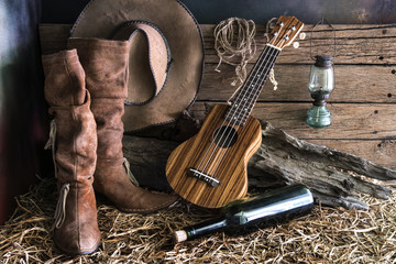 Still life photography with ukulele on american west rodeo brown felt cowboy hat and traditional leather boots in vintage ranch barn background