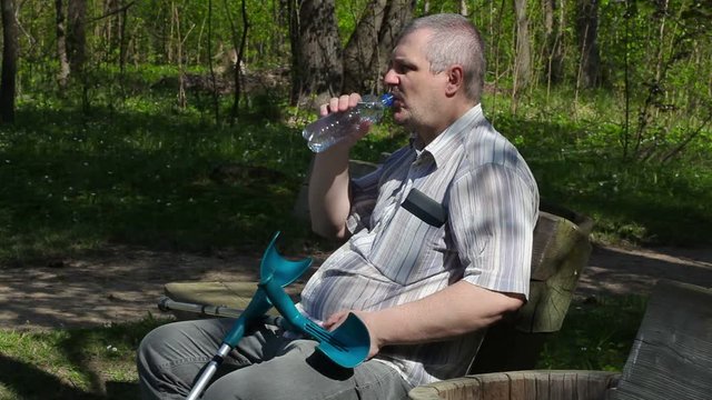 Disabled man with crutches sitting on the bench and drinking water