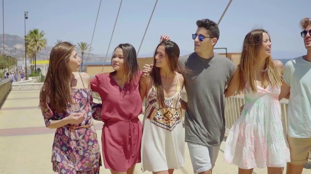Group of trendy young friends walking arm in arm along a seafront promenade smiling with pleasure as they enjoy their summer vacation