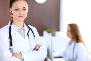 Beautiful young smiling female doctor standing in a hospital with her colleague in the background