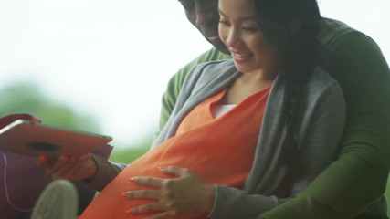  Happy couple expecting a baby relaxing in the park with computer tablet - Powered by Adobe