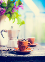 Espresso cup full of coffee beans on kitchen table with coffee pot on background of terrace,sky and flowers