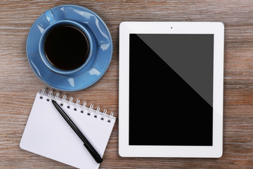 Tablet with cup of coffee and notepad on wooden table