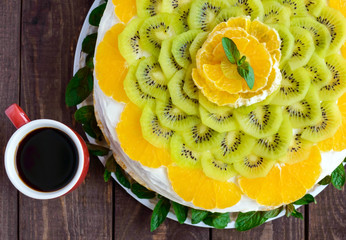 Bright round festive fruit cake decorated with kiwi, orange, mint and a cup of coffee.