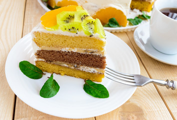 A piece of fruit cake (kiwi, orange, mint leaves) on a white plate on wooden background close-up and a cup of tea