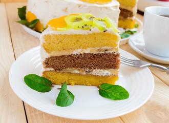 A piece of fruit cake (kiwi, orange, mint leaves) on a white plate on wooden background close-up and a cup of tea