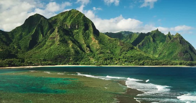 Aerial view flying over tropical coral reef and breaking ocean waves towards beautiful green mountains on Kauai