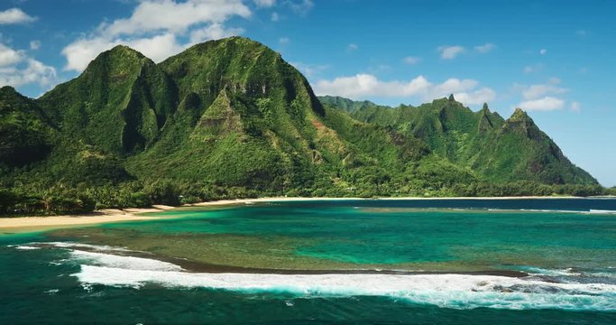 Aerial view flying over tropical coral reef and breaking ocean waves towards beautiful green mountains on Kauai
