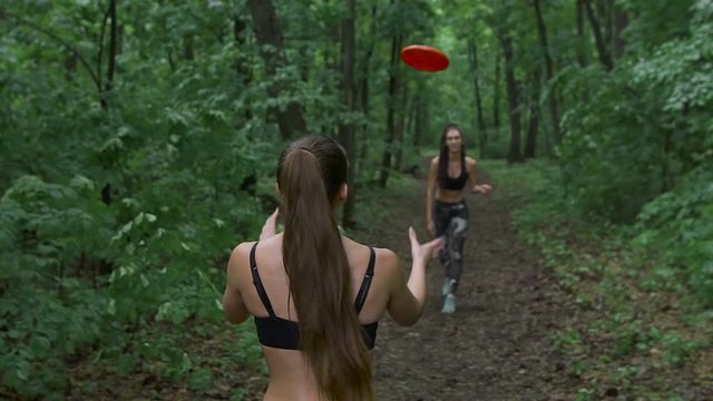 Two slender brunette in the woods, throwing a frisbee plate. They are having fun and playing on the footpath. 