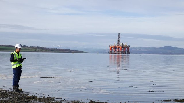 An asian engineer standing in front of semi submersible oil rig with a clipboard in Invergordon Scotland

