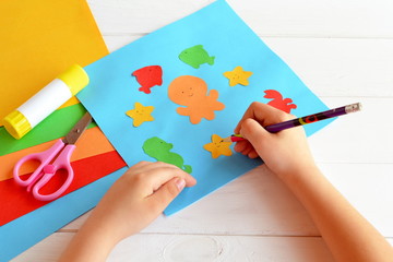 Child holds a pencil and draws. Child doing a card with sea animals and fish. Sheets of colored paper, scissors, glue, set for kids art 