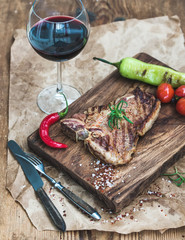 Cooked meat t-bone steak on serving board with roasted tomatoes, chili peppers, fresh rosemary, spices and glass of red wine over rustic wooden background