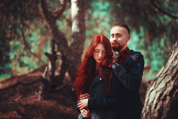 Fototapeta premium The red-haired guy with a beard and curly red-haired girl on the background of fabulous scenery of nature. Beautiful loving couple on a walk in the woods.