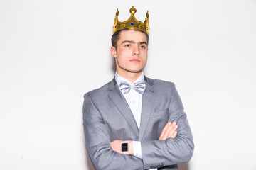 Sneering young handsome man wearing suit and crown keeping arms crossed and looking at camera while standing against white background