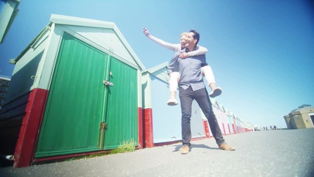  Couple playing piggyback pause for a selfie in front of colourful beach huts