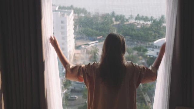 Young woman open curtains of hotel resort in tropical island, high flor, 4k uhd 2160p