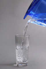 Filter jug pouring water in the glass, on the grey background, close-up