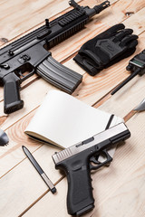 Assault rifle, gun, knife with sheath, compass and notebook with pen on table.