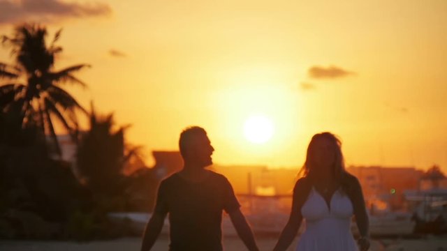 caucasian couple walking on the beach at sunset talking holding hands backlit