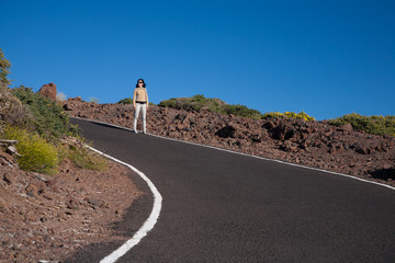 brunette woman brown jacket white trousers standing in curve of rural grey road with blue sky of La Palma, Canary Islands, Spain Europe