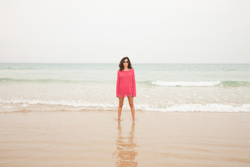 Fototapeta na wymiar brunette summer vacation woman with sunglasses, red sweater blue jeans shorts barefoot standing at seashore on sand with ocean behind, in beach in Cadiz, Andalusia, Spain, Europe 