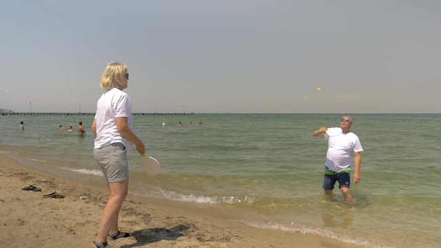 Slow motion shot of mature man and woman playing beach tennis, man is standing in the sea water, woman is on the sand.