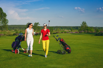 Two smiling sportive women golfers walking on golf course at sunny day