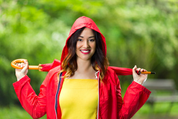 Portrait of a beautiful young woman in hooded red raincoat holding red umbrella. She is looking at camera and smiling while walking in the park on a rainy day.