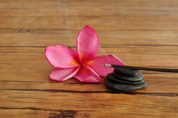 A pink frangipani flower displayed with a stack of pebbles and burning incense sticks