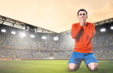 unhappy soccer or football player kneeled down with hands on his face at stadium