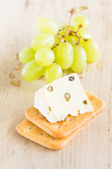 Eine Scheibe Weichkäse mit grünem Pfeffer liegt auf Kräckern. Im Hintergrund liegen Weintrauben.