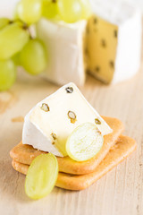 Eine Scheibe Weichkäse mit grünem Pfeffer liegt auf Kräckern. Im Hintergrund liegen ein angeschnittener Weichkäse und Weintrauben.