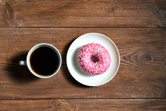Cup of black coffee with pink donut on a plate on wooden table, top view