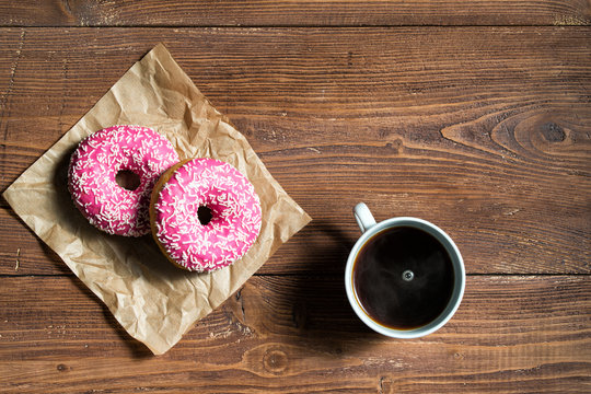 Cup of black coffee with two pink donuts on wooden background, top view